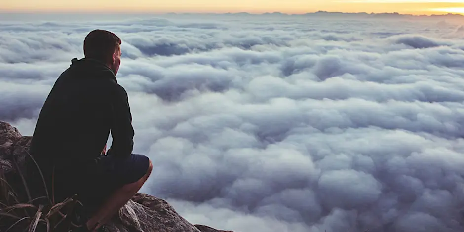 Photo of a man crouching on a mountain peak. He's looking down on clouds as the son rises.